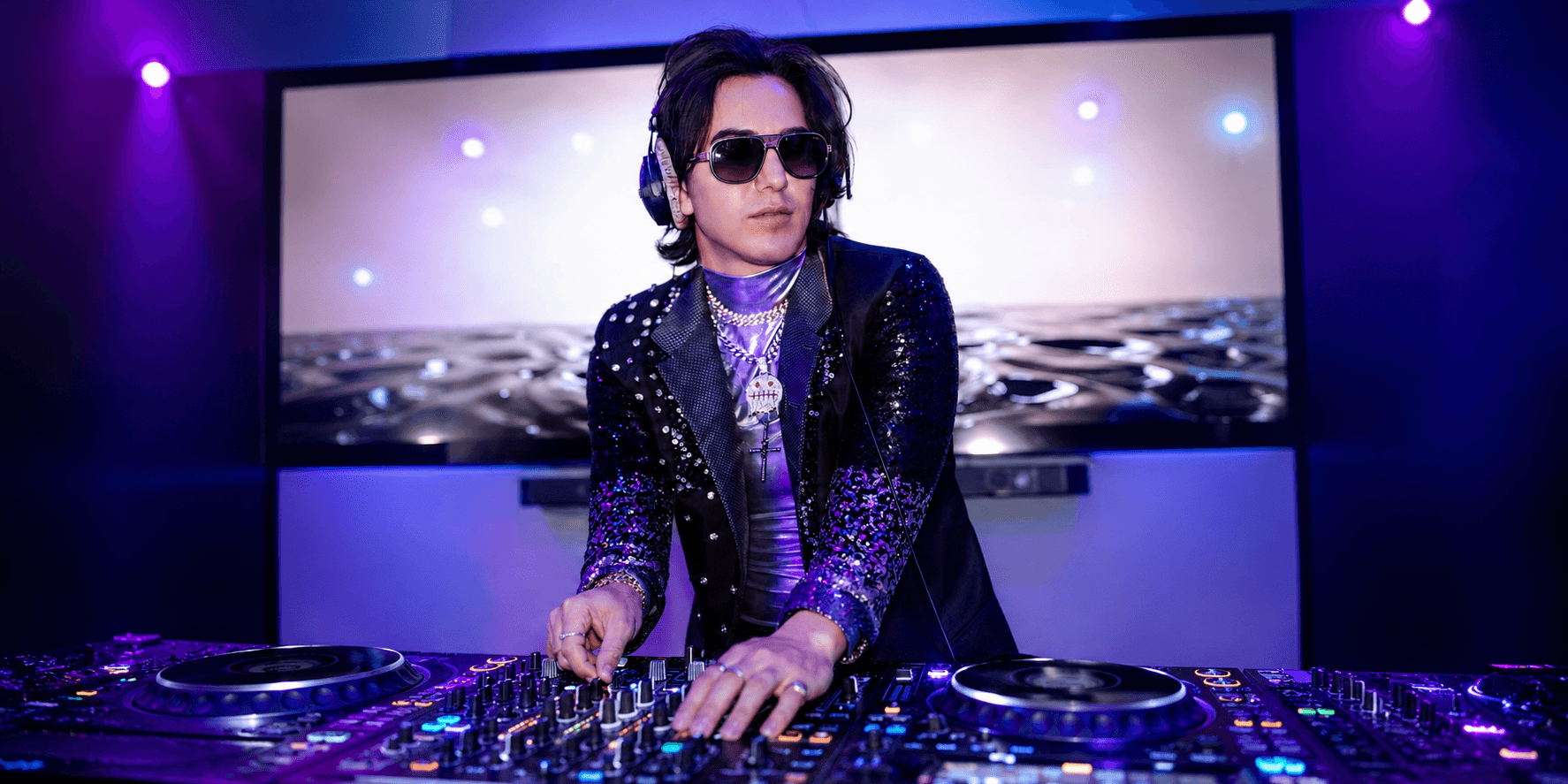 A DJ in sunglasses performs on turntables under purple lights with a digital backdrop