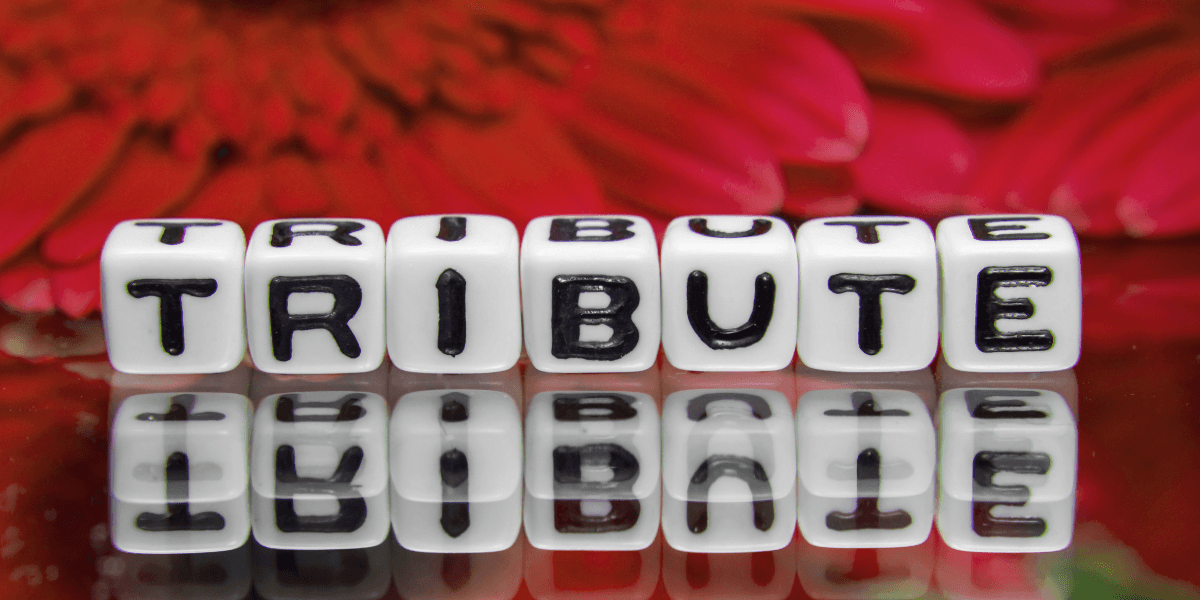White dice with the word "TRIBUTE" printed, reflecting on a shiny surface with flowers in background