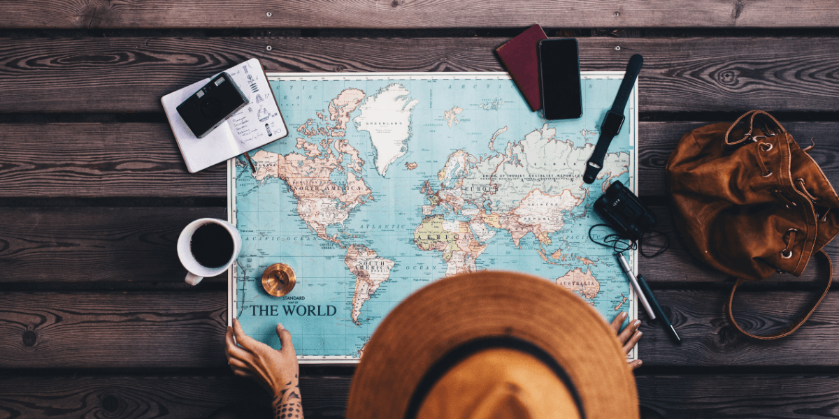 A person in a hat studies a world map on a wooden table with travel items.