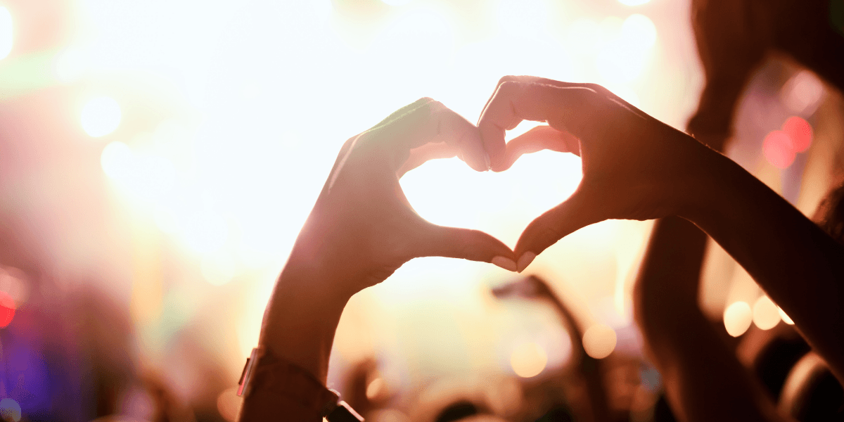 Hands forming a heart shape against a vibrant concert background with blurred lights