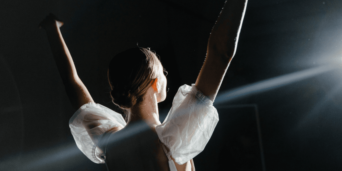 A dancer with their back turned raises arms, illuminated by spotlight in a dark space