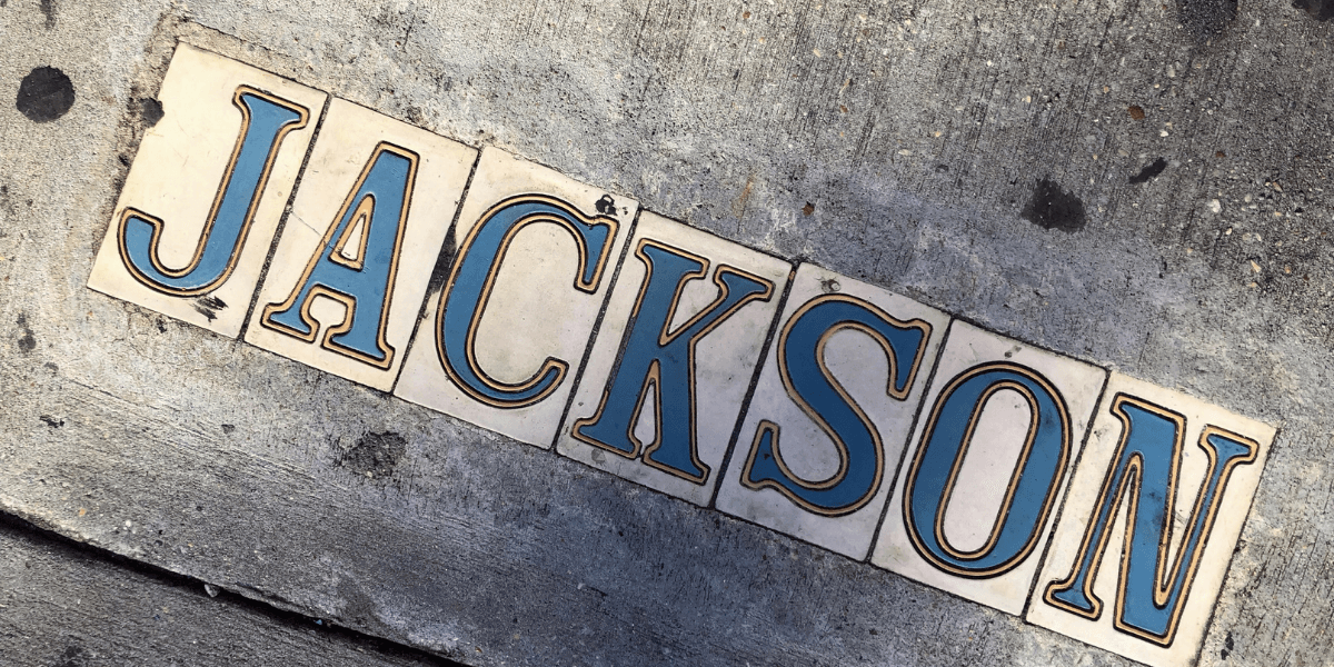 Decorative sidewalk tiles spelling “JACKSON” embedded in a concrete pavement.