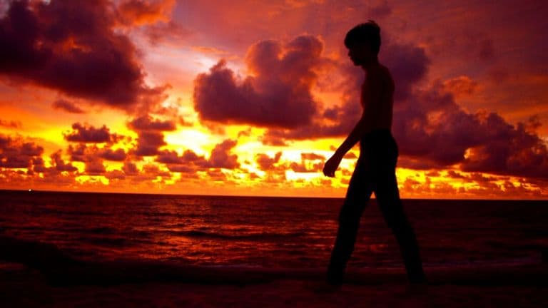 a silhouette of a person walking along a beach during a vibrant sunset, with dramatic clouds in the sky.