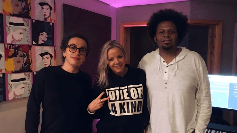 Three people pose in music studio with pop-art portraits on wall and glowing computer monitor.