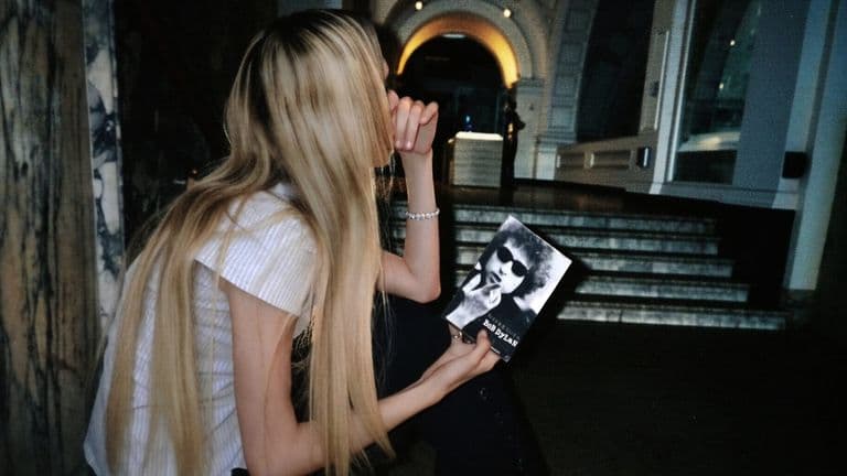Blonde woman holding Bob Dylan book inside dimly lit building with dramatic architectural background.