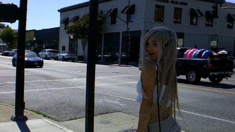 Smiling person with long white hair and angel wings standing on sunny city sidewalk.