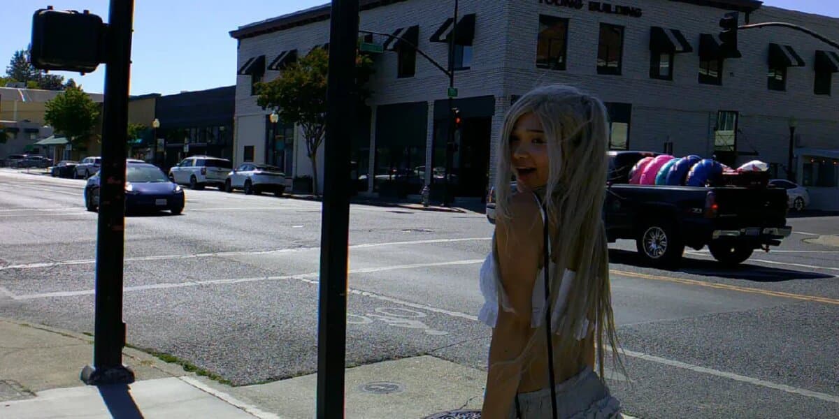 Smiling person with long white hair and angel wings standing on sunny city sidewalk.