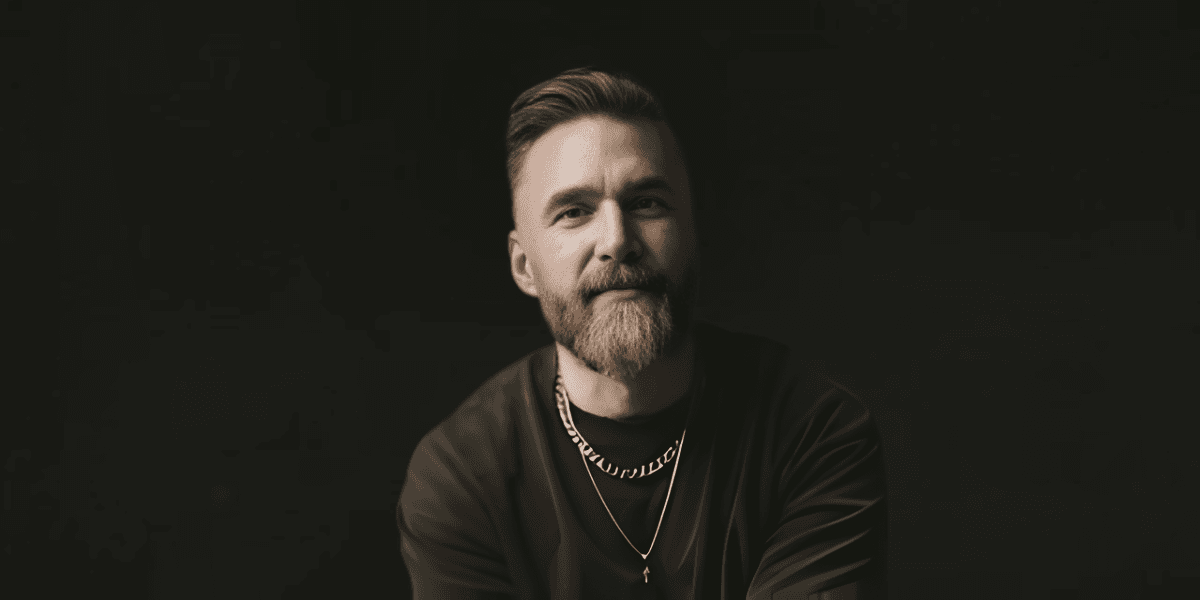 Portrait of a bearded man wearing necklaces against a dark background, looking calmly toward the camera.