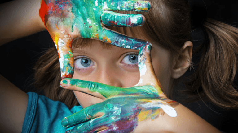 Close-up of a young girl with blue eyes, playfully framing her face with colorful painted hands, showcasing vibrant shades of green, yellow, and red paint. The image highlights her creativity and love for art against a dark background.