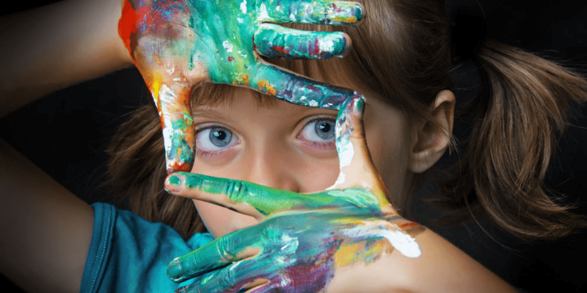 Close-up of a young girl with blue eyes, playfully framing her face with colorful painted hands, showcasing vibrant shades of green, yellow, and red paint. The image highlights her creativity and love for art against a dark background.