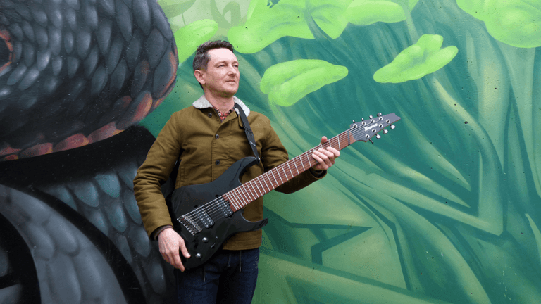 A musician standing outdoors, holding a black electric guitar, with a vibrant green and black graffiti wall in the background. The musician is wearing a brown jacket, denim jeans, and a confident expression, creating an artistic and dynamic look.