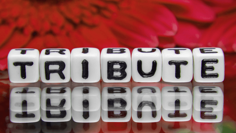 White dice with the word "TRIBUTE" printed, reflecting on a shiny surface with flowers in background