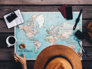 A person in a hat studies a world map on a wooden table with travel items.