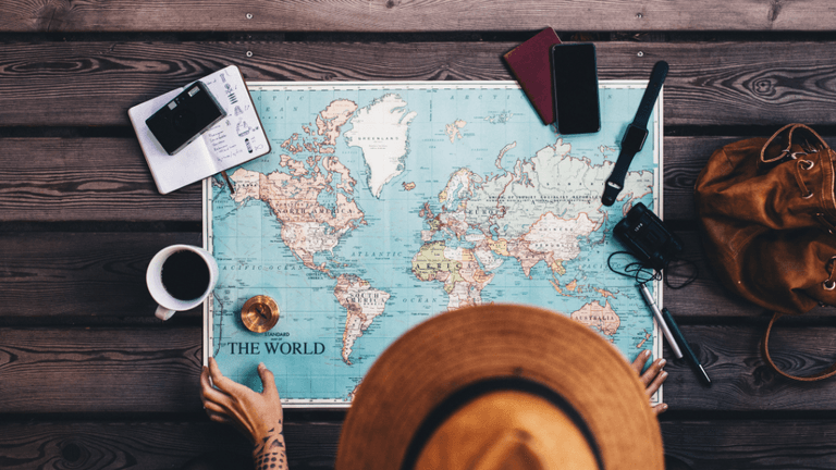 A person in a hat studies a world map on a wooden table with travel items.