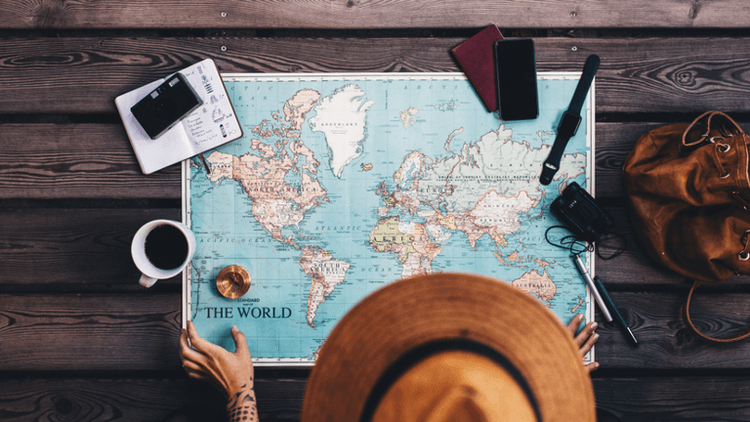 A person in a hat studies a world map on a wooden table with travel items.