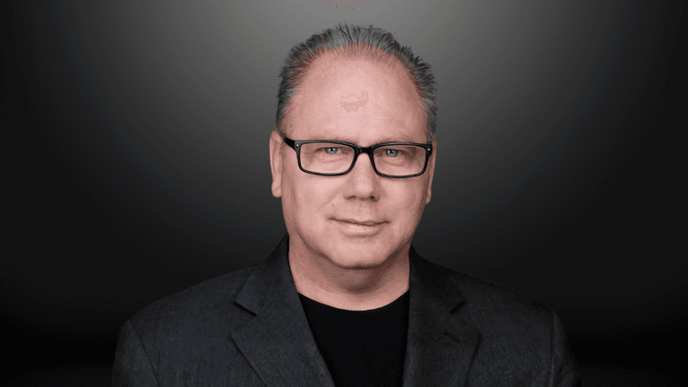 Middle-aged man with glasses and gray hair in a dark blazer against a gray background