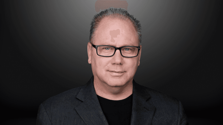 Middle-aged man with glasses and gray hair in a dark blazer against a gray background