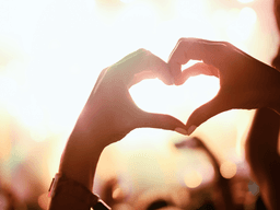 Hands forming a heart shape against a vibrant concert background with blurred lights