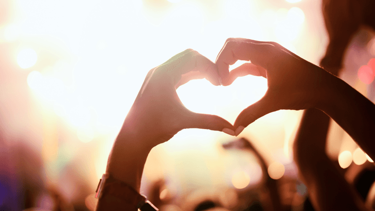Hands forming a heart shape against a vibrant concert background with blurred lights