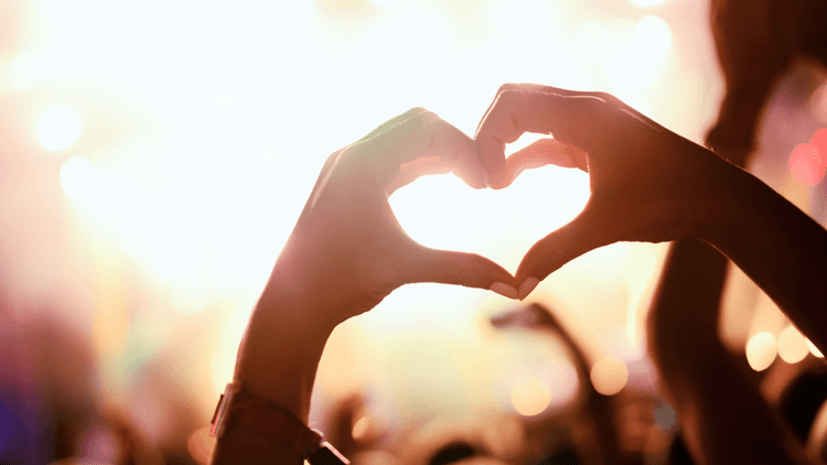 Hands forming a heart shape against a vibrant concert background with blurred lights