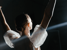 A dancer with their back turned raises arms, illuminated by spotlight in a dark space