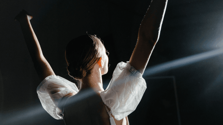 A dancer with their back turned raises arms, illuminated by spotlight in a dark space