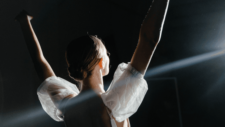 A dancer with their back turned raises arms, illuminated by spotlight in a dark space