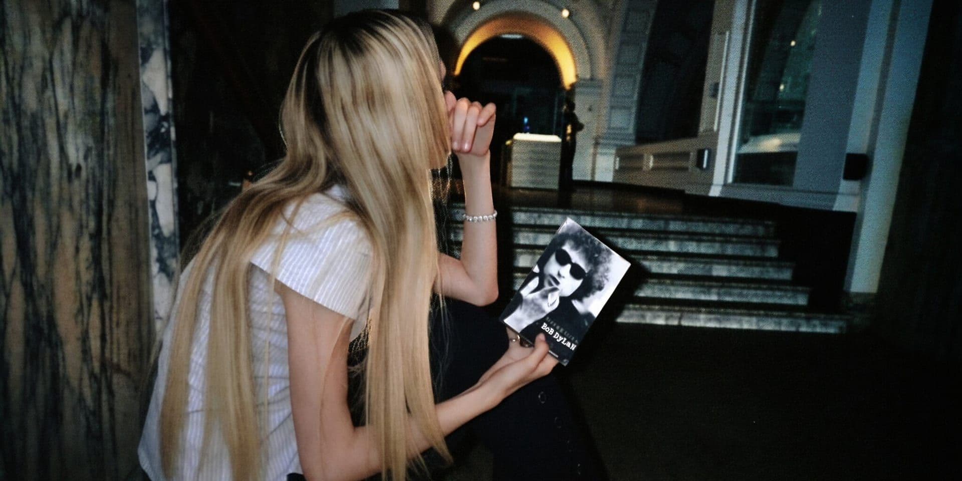 Blonde woman holding Bob Dylan book inside dimly lit building with dramatic architectural background.