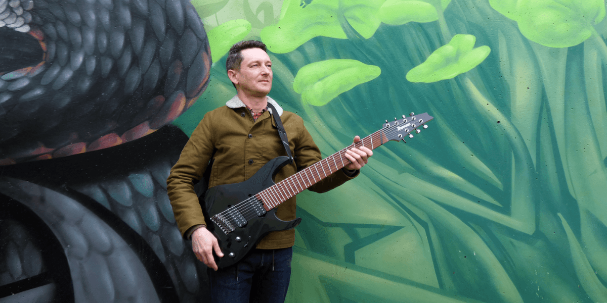 A musician standing outdoors, holding a black electric guitar, with a vibrant green and black graffiti wall in the background. The musician is wearing a brown jacket, denim jeans, and a confident expression, creating an artistic and dynamic look.
