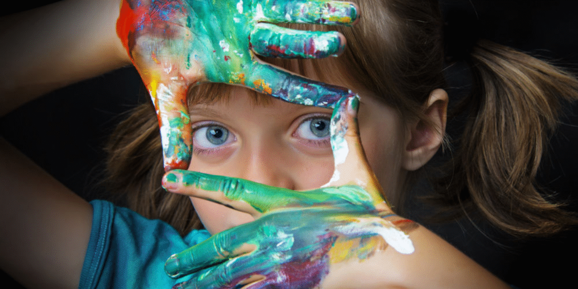 Close-up of a young girl with blue eyes, playfully framing her face with colorful painted hands, showcasing vibrant shades of green, yellow, and red paint. The image highlights her creativity and love for art against a dark background.