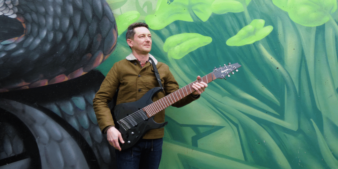 A musician standing outdoors, holding a black electric guitar, with a vibrant green and black graffiti wall in the background. The musician is wearing a brown jacket, denim jeans, and a confident expression, creating an artistic and dynamic look.