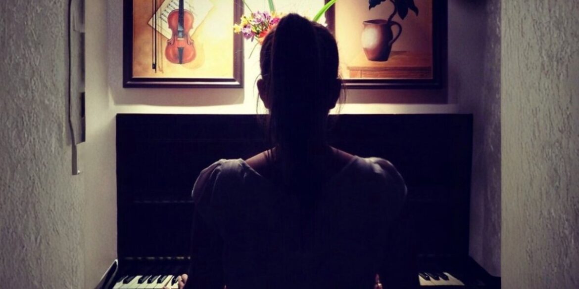 A woman sits at a piano, with her back to the camera, framed by art pieces and soft, ambient light.