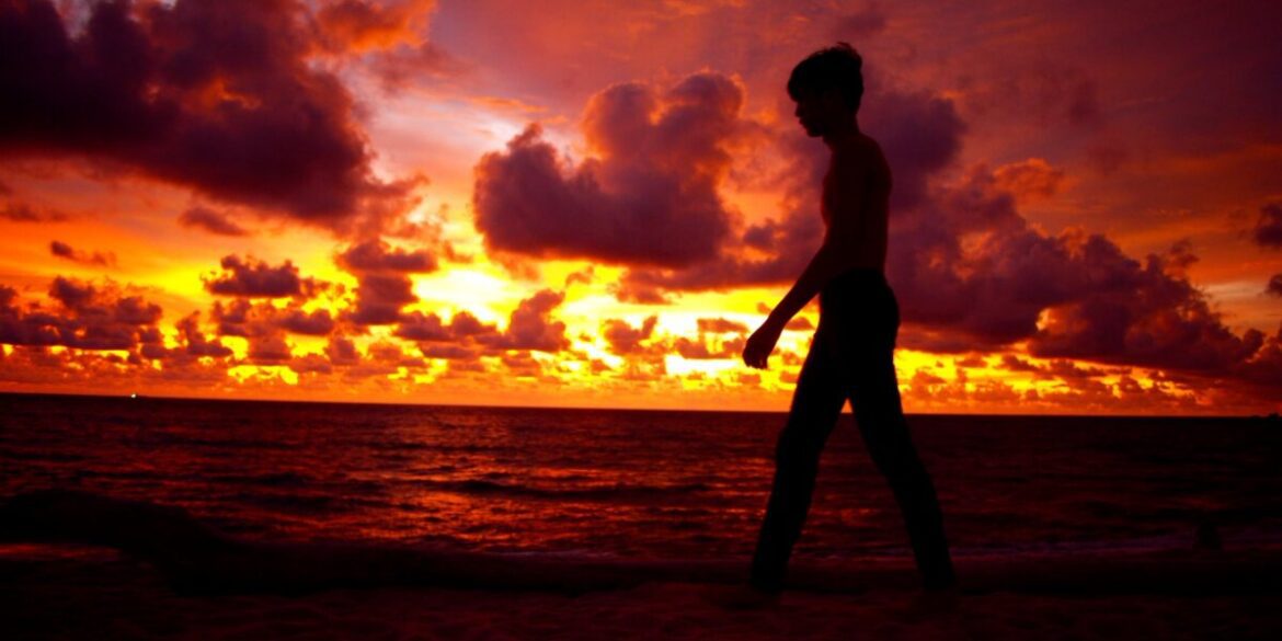 a silhouette of a person walking along a beach during a vibrant sunset, with dramatic clouds in the sky.