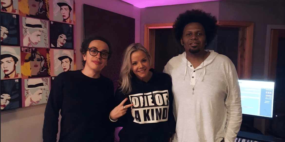 Three people pose in music studio with pop-art portraits on wall and glowing computer monitor.