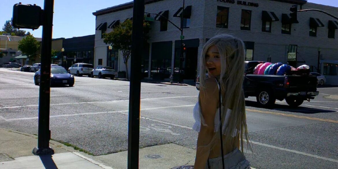 Smiling person with long white hair and angel wings standing on sunny city sidewalk.